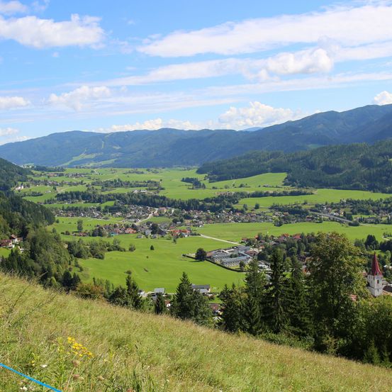 Ein Panoramablick auf ein Tal mit üppigen grünen Feldern, verstreuten Häusern und einem Kirchturm in der Ferne, alles unter einem blauen Himmel mit Wolken.
