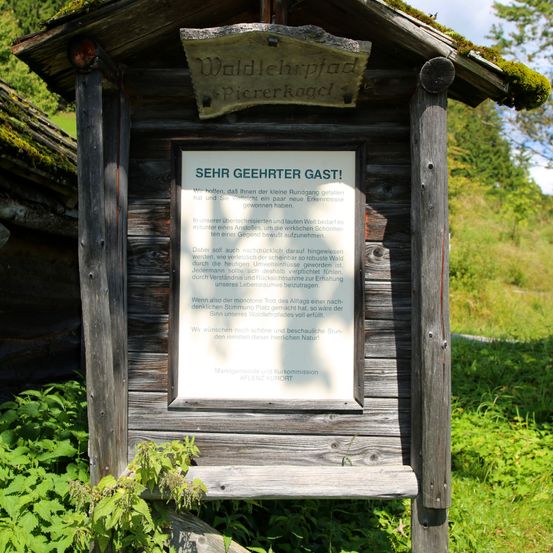 Ein hölzernes Schild mit grünem Dach in einem Wald trägt die Aufschrift 'SEHR GEEHRTER GAST!' mit zusätzlichem Text und einem Logo. Es ist von Pflanzen und Bäumen umgeben.