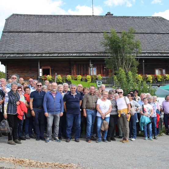Eine große Gruppe von Menschen posiert für ein Foto vor einem Holzhaus mit Balkon und vielen Blumen.