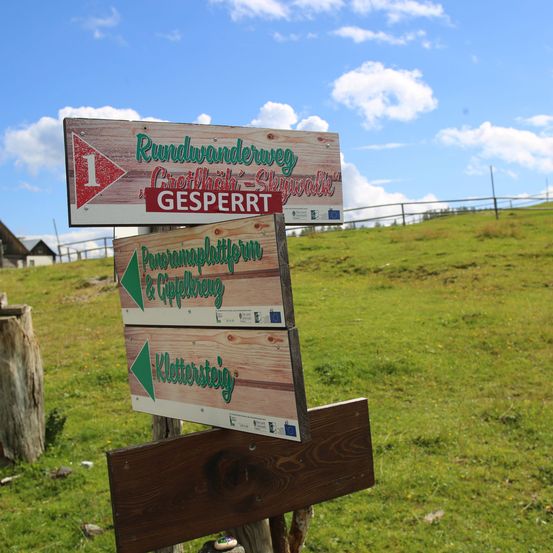 Drei Holzschilder auf einem Pfosten weisen auf Wege hin: Rundwanderweg, Panoramaplattform & Gipfelkreuzung und Klettersteig. Das erste Schild ist mit 1 nummeriert. Das Gebiet ist grasig mit einem Zaun und einem Haus im Hintergrund.