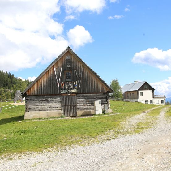 Eine Holzhütte mit einem dreieckigen Dach und einer Holztür steht auf einer Kiesstraße. An der Wand hängen Skier und andere Gegenstände. Dahinter steht ein kleines Haus. Der Himmel ist klar und blau.