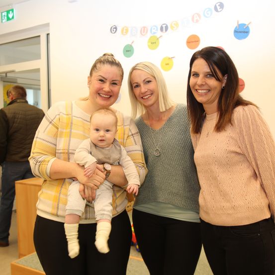 Drei Frauen und ein Baby lächeln in einem Raum. Die Frau auf der linken Seite hält das Baby. Hinter ihnen ist eine Wand mit einem farbenfrohen Banner zu sehen, auf dem 'Geburtstag' steht.