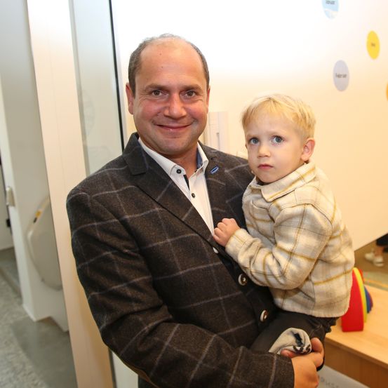 Ein Mann in einem karierten Blazer lächelt, während er einen jungen Jungen mit blonden Haaren hält. Der Junge sieht ernst aus. Sie stehen in einem Raum mit einer bunten Wand.