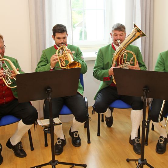 Vier Musiker in grünen Uniformen sitzen auf blauen Stühlen und spielen Trompeten und Tuba, mit Notenständern vor ihnen und einem Fenster dahinter.