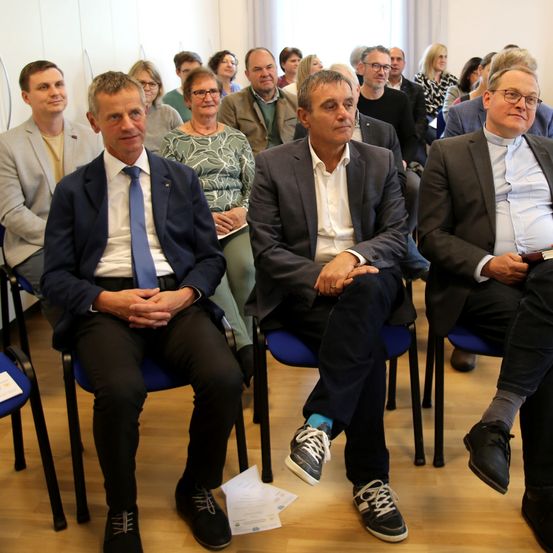 Eine Gruppe von Menschen in formeller Kleidung sitzt in einer Reihe blauer Stühle in einem Besprechungsraum.