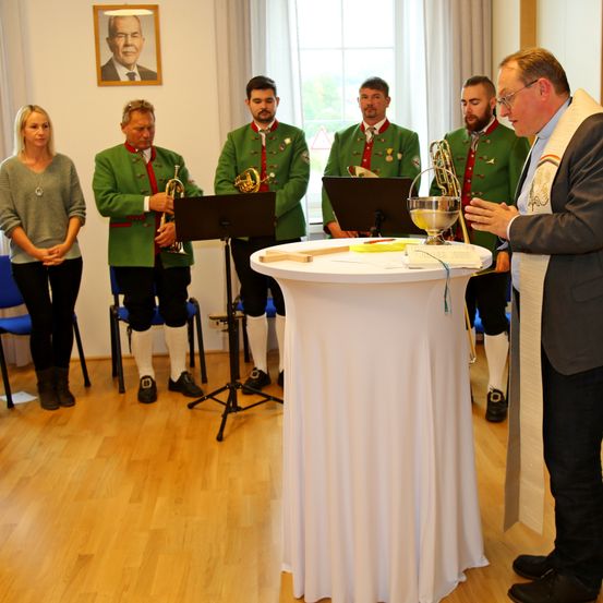 Ein Mann spricht vor einem Tisch mit einem Kelch, während Musiker in grünen Uniformen hinter ihm stehen. Eine Frau steht links. Ein Porträt ist an der Wand.