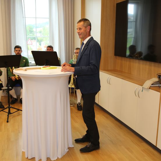 Ein Mann in einem Anzug steht an einem Rednerpult mit einem Mikrofon und spricht andere an. Hinter ihm sitzen Musiker mit Trompeten und Posaunen. Ein Monitor ist an der Wand montiert und ein Fenster blickt nach draußen.