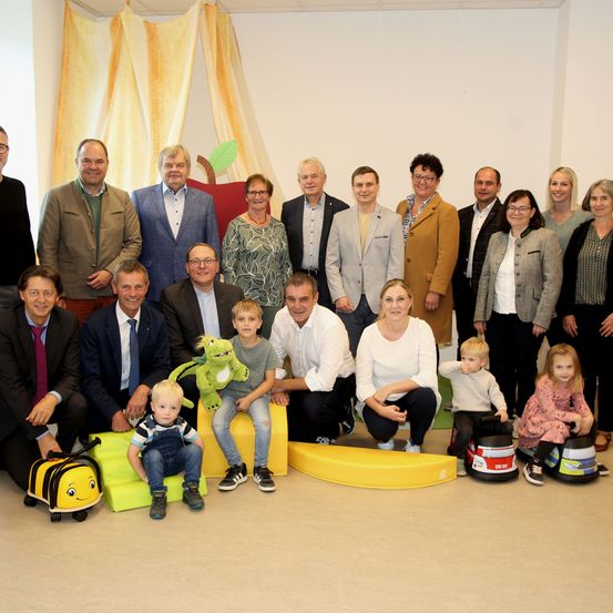 Eine Gruppe von Erwachsenen und Kindern posiert für ein Foto in einem Raum mit weißer Wand und gelben Möbeln. Einige Kinder sitzen auf gelben Stühlen, andere stehen. Ein erwachsener Mann hält einen grünen Spielzeugfrosch.
