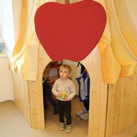 Ein kleiner Junge steht in einem holzverkleideten Apfelspielhaus und hält einen gelben Becher. Er ist von einem gelben Vorhang und zwei Personen in Turnschuhen umgeben.
