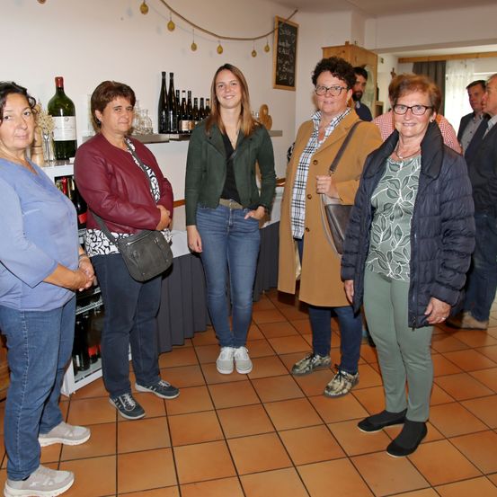 Fünf Frauen stehen in einem Raum mit gefliestem Boden, wahrscheinlich bei einer Weinverkostung. Sie alle lächeln und schauen nach vorne. Hinter ihnen befindet sich ein Regal mit Weinflaschen und einer Tafel.