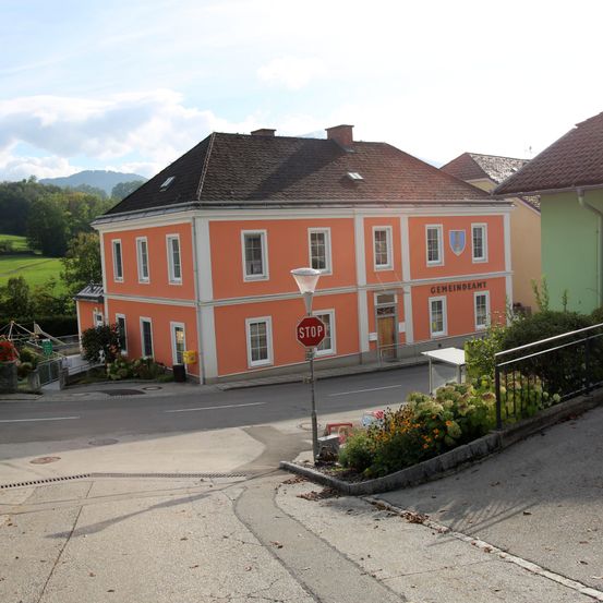 Eine Straßenszene mit einem orangefarbenen Gebäude mit der Aufschrift 'Gemeindeamt', einem Stoppschild und einem grünen Feld im Hintergrund.