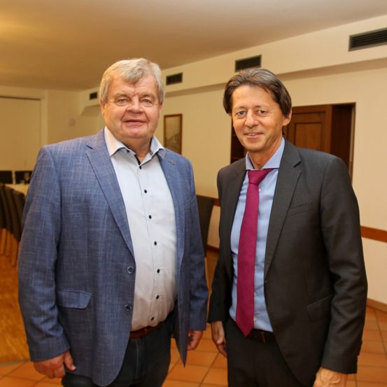 Zwei Männer stehen nebeneinander in einem Raum und lächeln. Der Mann auf der linken Seite trägt einen blauen Anzug mit einem weißen Hemd und einem braunen Gürtel. Der Mann auf der rechten Seite trägt einen schwarzen Anzug mit einer roten Krawatte. Hinter ihnen ist eine Holztür, und im Hintergrund befindet sich ein Tisch mit Stühlen.