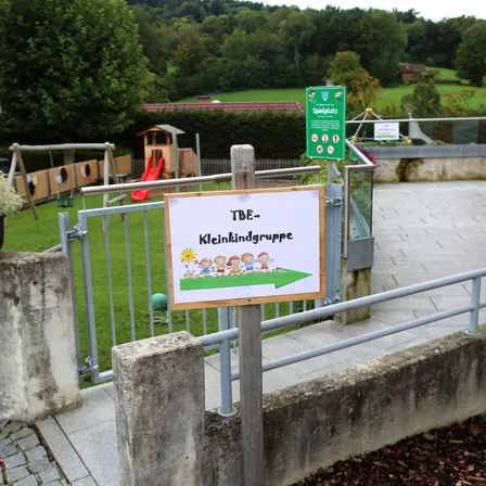 Ein Spielplatz mit einer roten Rutsche, einer Schaukel und einem kleinen Holzhaus ist eingezäunt. Ein Schild weist auf einen Eingang für eine Kleinkindgruppe hin.