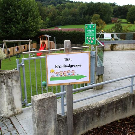 Ein Spielplatz mit einer roten Rutsche, einer Schaukel und einem kleinen Holzhaus ist eingezäunt. Ein Schild weist auf einen Eingang für eine Kleinkindgruppe hin.