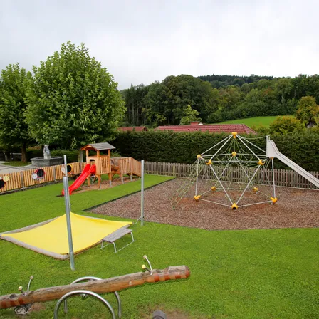 Freiluftspielplatz mit Rutsche, Klettergerüst und einem großen Baum. Der Bereich ist von einem Holzzaun umgeben und hat grünen Rasen.