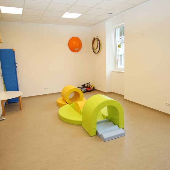 Ein Spielzimmer mit hellbraunem Boden hat bunte Spielgeräte. Ein blaues Kissen und ein runder orangefarbener Ballon sind an der Wand. Ein Fenster befindet sich an der rechten Wand.