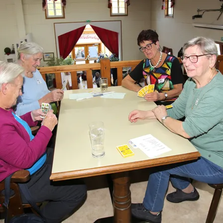 Vier ältere Frauen sitzen um einen Tisch herum und spielen ein Kartenspiel. Sie lächeln und scheinen sich zu amüsieren.