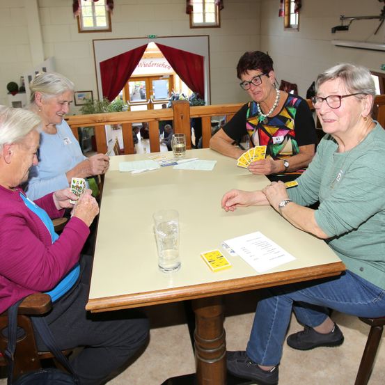Vier ältere Frauen sitzen um einen Tisch herum und spielen ein Kartenspiel. Sie lächeln und scheinen sich zu amüsieren.