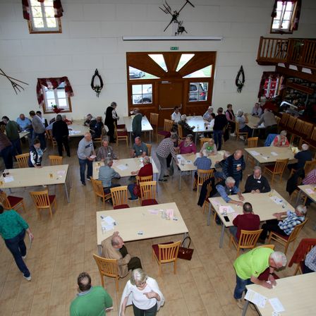 Ein Raum mit mehreren Tischen und Stühlen ist mit Menschen gefüllt. Sie scheinen an einer Aktivität beteiligt zu sein, vielleicht eine Besprechung oder eine Veranstaltung. Einige haben Papiere auf ihren Tischen.