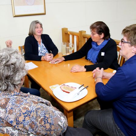 Fünf Frauen sitzen um einen Holztisch und führen ein Gespräch. Ein Teller mit einem Sandwich und eine Gabel steht auf dem Tisch. Jede Frau trägt eine Brille, und eine hat eine Schürze.