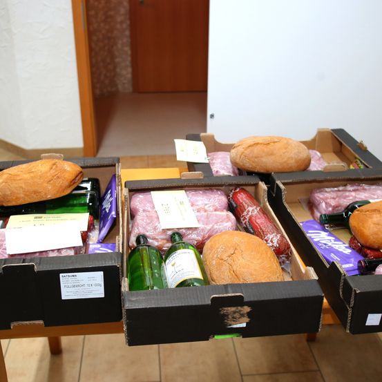 Vier Kisten auf einem Tisch enthalten verschiedene Artikel: zwei Brote, zwei Flaschen Schnaps, Würste und eine Schokoladentafel. Die Kisten sind mit Produktnamen und Gewichten beschriftet.