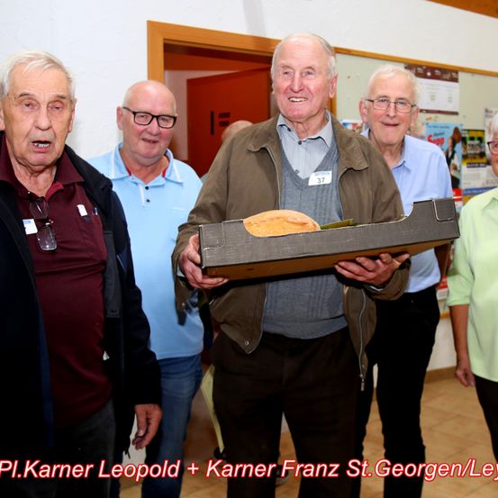 Fünf ältere Männer stehen zusammen, einer hält eine Schachtel mit Brot darin. Sie lächeln und scheinen für ein Foto zu posieren.