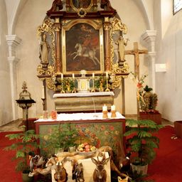 Ein kunstvoll verzierter Altar mit Gemälde, Kerzen und einem Kreuz steht in einer Kapelle. Verschiedene Pflanzen, Früchte und Tierköpfe schmücken die Vorderseite des Altars.