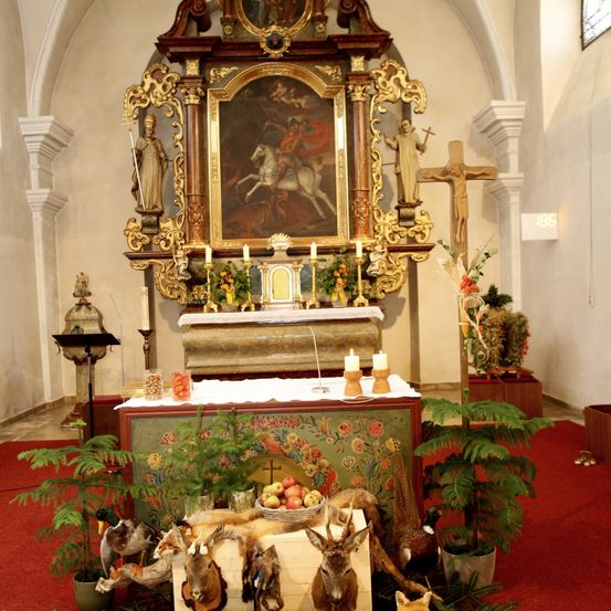 Ein kunstvoll verzierter Altar mit Gemälde, Kerzen und einem Kreuz steht in einer Kapelle. Verschiedene Pflanzen, Früchte und Tierköpfe schmücken die Vorderseite des Altars.