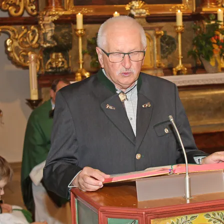 Ein älterer Mann steht an einem Podium und liest aus einem Buch, trägt einen formellen Anzug und eine Brille. Hinter ihm befindet sich ein Altar mit Kerzen und einer Blumenarrangement. Eine andere Person ist teilweise im Hintergrund sichtbar.