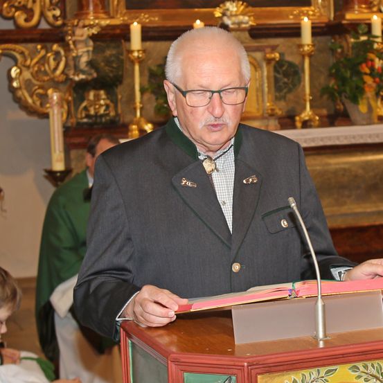 Ein älterer Mann steht an einem Podium und liest aus einem Buch, trägt einen formellen Anzug und eine Brille. Hinter ihm befindet sich ein Altar mit Kerzen und einer Blumenarrangement. Eine andere Person ist teilweise im Hintergrund sichtbar.