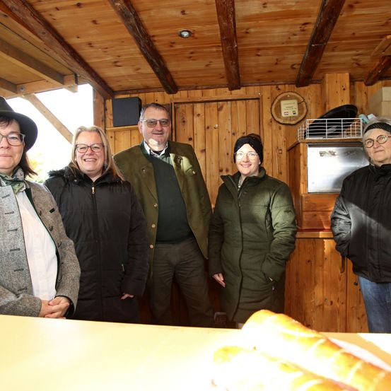 Fünf Personen stehen lächelnd und posieren für ein Foto in einer Holzhütte. Ein Tisch mit Brot steht im Vordergrund. Die Hütte hat Holzwände und eine Holzdecke.