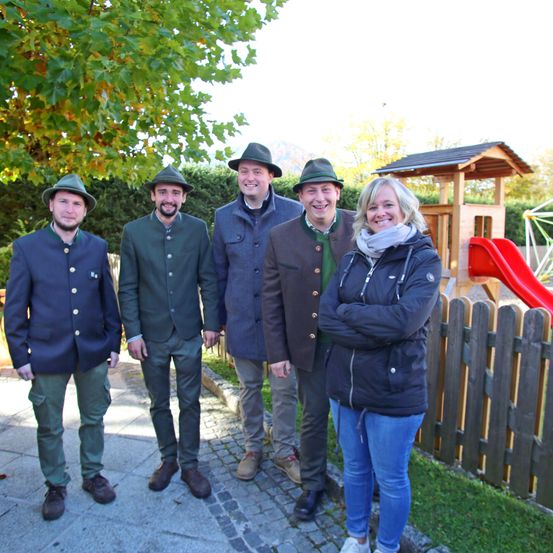 Fünf Personen stehen in einem Park mit einem Spielplatz hinter ihnen. Sie alle lächeln. Vier von ihnen tragen Hüte.