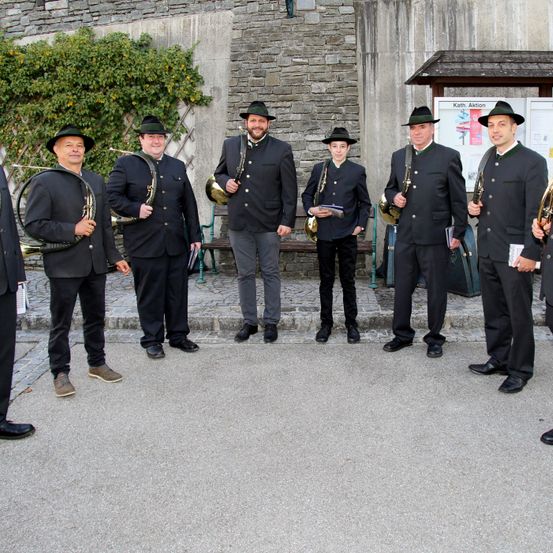 Eine Gruppe von Männern in formeller Kleidung, die Musikinstrumente halten, steht vor einer Steinmauer mit Grünpflanzen und einer Holzbank.