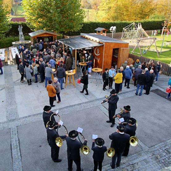 Eine Freiluftveranstaltung, bei der eine Gruppe von Menschen steht, einige sitzen an Tischen. Eine Band spielt in der Mitte. Es gibt auch einige Kinder, die im Spielbereich spielen.