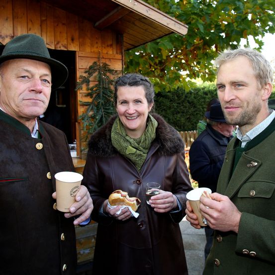 Drei Menschen stehen draußen in der Nähe eines Holzgebäudes und lächeln für ein Foto. Der Mann auf der linken Seite hält einen Becher und ein Sandwich, die Frau in der Mitte hält ein Sandwich und ein Glas und der Mann auf der rechten Seite hält einen Becher. Hinter ihnen steht ein weiterer Mann in der Nähe eines Busches.