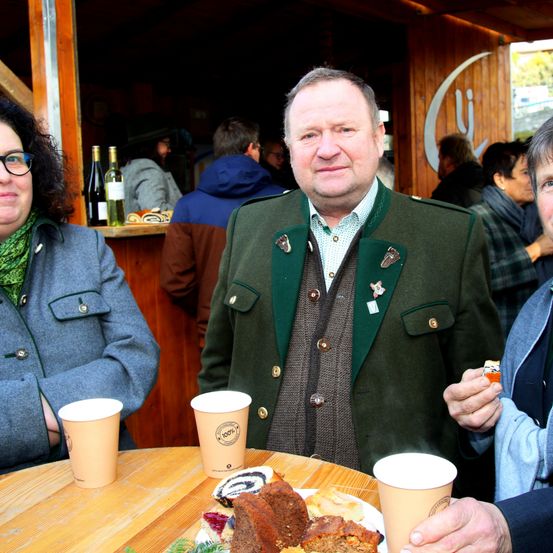 Ein Mann und zwei Frauen sitzen an einem Tisch in einer rustikalen Umgebung, mit Essen und Getränken vor ihnen. Der Mann trägt eine grüne Jacke mit Knöpfen und einer Abzeichen. Dahinter zeigt eine verschwommene Szene Menschen im Stehen und eine Holzwand mit einem Logo.