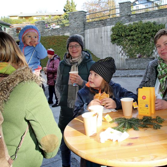 Eine Gruppe von Menschen versammelt sich um einen Tisch, möglicherweise in einem Park. Ein junges Mädchen isst ein Sandwich, während eine Frau einen Becher hält. Hinter ihnen stehen eine Frau und ein Kind. Ein gelbes Päckchen liegt auf dem Tisch.