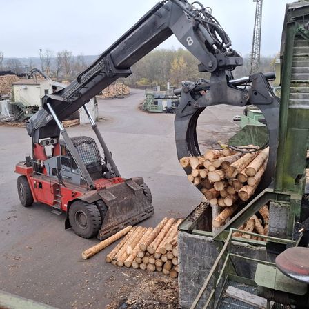 Ein roter Gabelstapler mit der Nummer 8 auf ihm hebt Holzstämme in einem Lagerhaus. Es gibt viele Holzstämme auf dem Boden und eine Holzpalette in der Nähe.