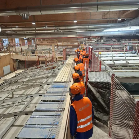 Eine Gruppe von Personen in Sicherheitswesten und Helmen steht auf einer Baustelle und inspiziert das Gebiet. Es gibt Metallgeländer und Absperrungen sowie einige Maschinen.