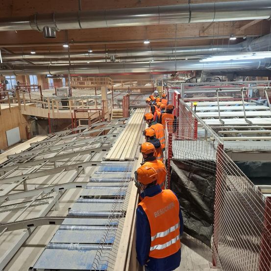Eine Gruppe von Personen in Sicherheitswesten und Helmen steht auf einer Baustelle und inspiziert das Gebiet. Es gibt Metallgeländer und Absperrungen sowie einige Maschinen.