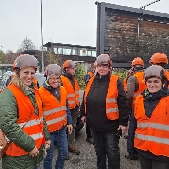 Eine Gruppe von Menschen in orangenen Westen und Helmen steht an einem bewölkten Tag im Freien. Ein Mann in einem schwarzen West steht in der Mitte. Hinter ihnen ist eine Holzwand mit einem Gebäude.
