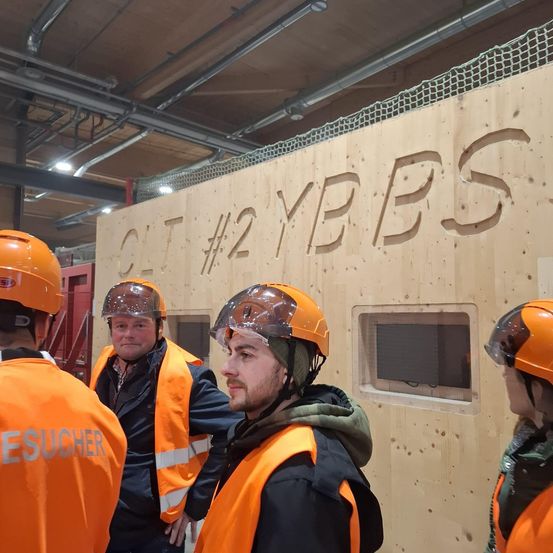 Vier Personen in orangefarbenen Westen und Helmen stehen vor einer Holzwand mit der Aufschrift 'CT #2 YBS'. Sie diskutieren wahrscheinlich arbeitsbezogene Angelegenheiten.