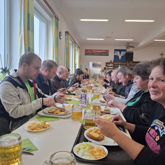 Eine Gruppe von Menschen sitzt an einem langen Tisch, isst Essen und trinkt Bier. Der Tisch ist mit Tellern voller Essen bedeckt, und vor jeder Person stehen Gläser mit Bier.