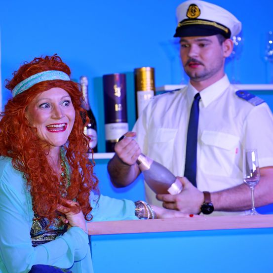 Eine Frau mit roten Haaren und einem Stirnband öffnet eine Champagnerflasche. Ein Mann in Uniform steht hinter ihr.