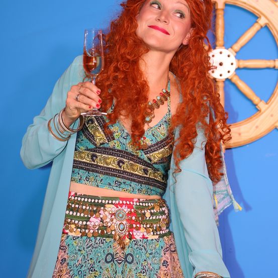 Eine Frau mit roten Haaren hält einen Weinglas vor einem blauen Hintergrund mit einem Lenkrad. Sie trägt ein farbenfrohes Kleid und eine Halskette.