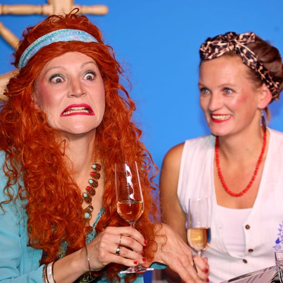 Zwei Frauen sitzen zusammen, eine in einem blauen Shirt, die ein Weinglas hält, die andere in einem weißen Shirt, lächelnd. Beide tragen Stirnbänder, eines mit Leopardenmuster. Sie scheinen für ein Foto zu posieren.
