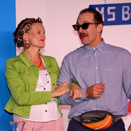 Ein Mann und eine Frau stehen eng beieinander, der Mann trägt eine Sonnenbrille und eine Bauchtasche, während die Frau eine grüne Jacke und rosa Hosen trägt. Sie scheinen in einem Studio mit einer rosa Wand und einem Schild zu stehen, das MS B liest.
