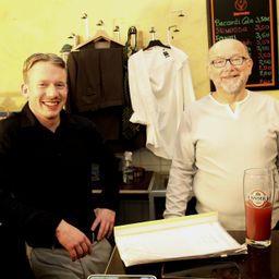 Zwei Männer sitzen vor einem Tisch. Der Mann auf der linken Seite lächelt, während der Mann auf der rechten Seite eine Brille trägt. Dahinter befindet sich ein Regal mit verschiedenen Gegenständen, und eine Tafel mit Preisen ist an der Wand montiert.