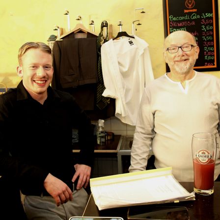 Zwei Männer sitzen vor einem Tisch. Der Mann auf der linken Seite lächelt, während der Mann auf der rechten Seite eine Brille trägt. Dahinter befindet sich ein Regal mit verschiedenen Gegenständen, und eine Tafel mit Preisen ist an der Wand montiert.