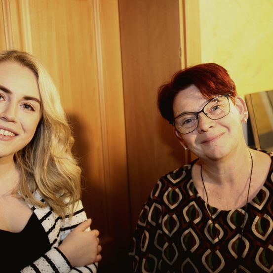 Zwei lächelnde Frauen, eine mit blonden Haaren und die andere mit kurzen roten Haaren, beide mit Brille. Sie stehen vor einer Holzwand.